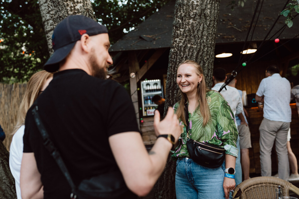 Two people are standing outside by the trees, talking and smiling. One person is wearing a green shirt and jeans, while the other is wearing a black shirt and a baseball cap. Other people and a wooden bar are visible in the background.  