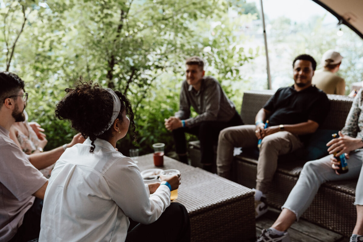 A group of people sit and chat on wicker furniture outdoors, surrounded by greenery. Some are holding drinks, and everyone seems relaxed and engaged in conversation. 