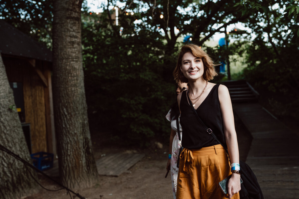 A smiling woman with short hair, wearing a black top and mustard yellow pants, stands outside on a forest path. She is holding a phone and carrying a crossbody bag, while trees and a wooden building can be seen in the background. 