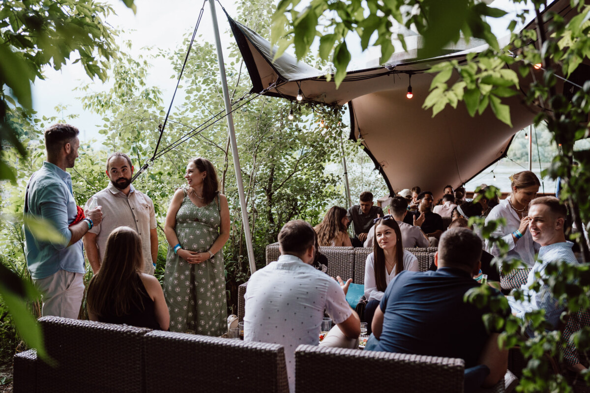 A group of people gather outside under a canopy surrounded by greenery, sitting and standing in conversation. The atmosphere seems relaxed and social, with a mix of men and women enjoying the event. 