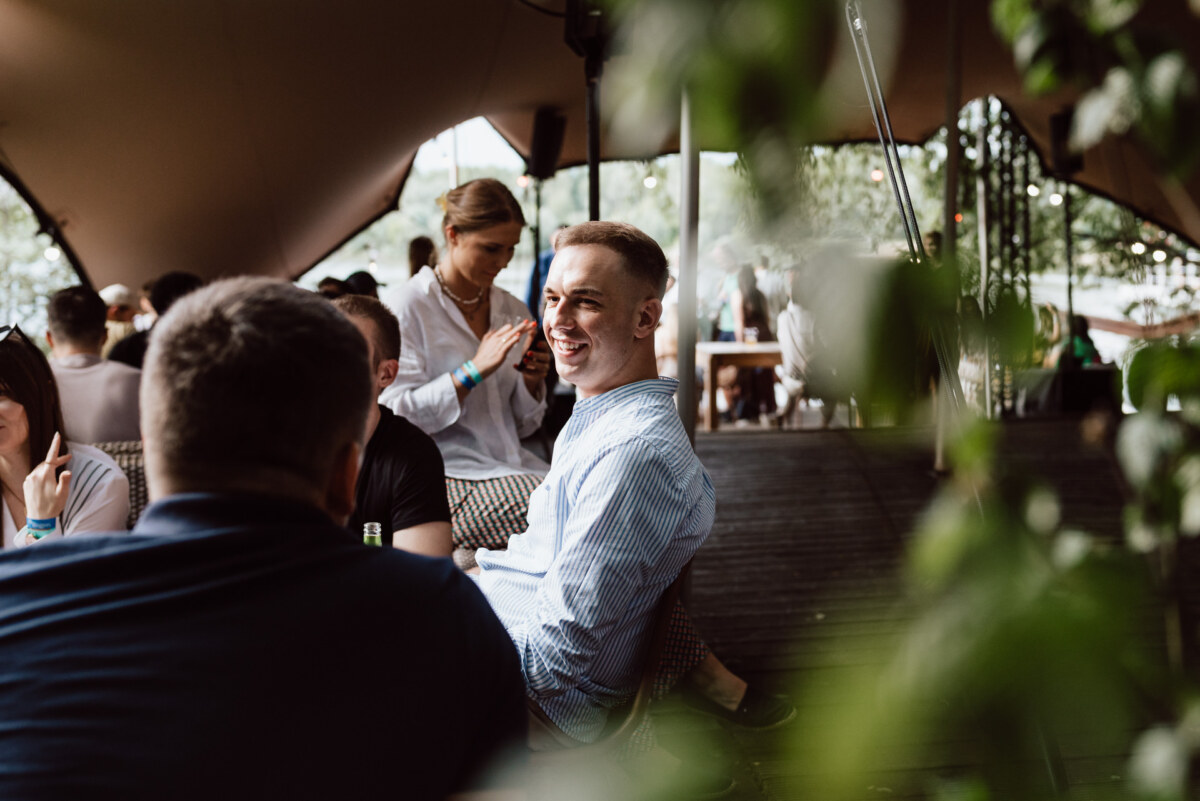 A group of people sit and talk at an outdoor party under a canopy. A man in a striped shirt smiles, surrounded by others, with greenery slightly blurred in the foreground. 
