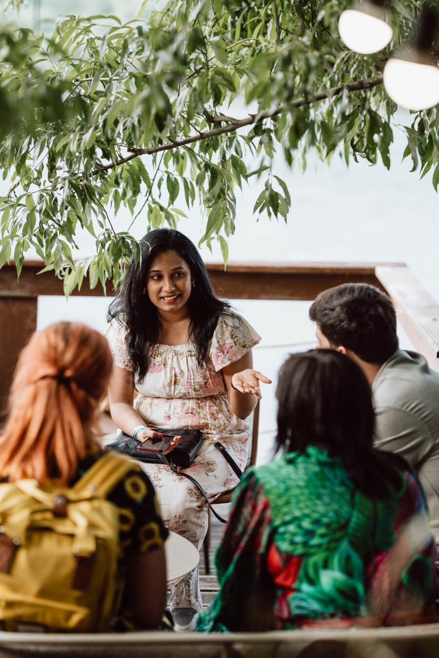 A woman in a flowery dress sits outside under leafy trees, talking and gesturing with her hand to three people who are listening intently. The group seems to be having a casual conversation. 