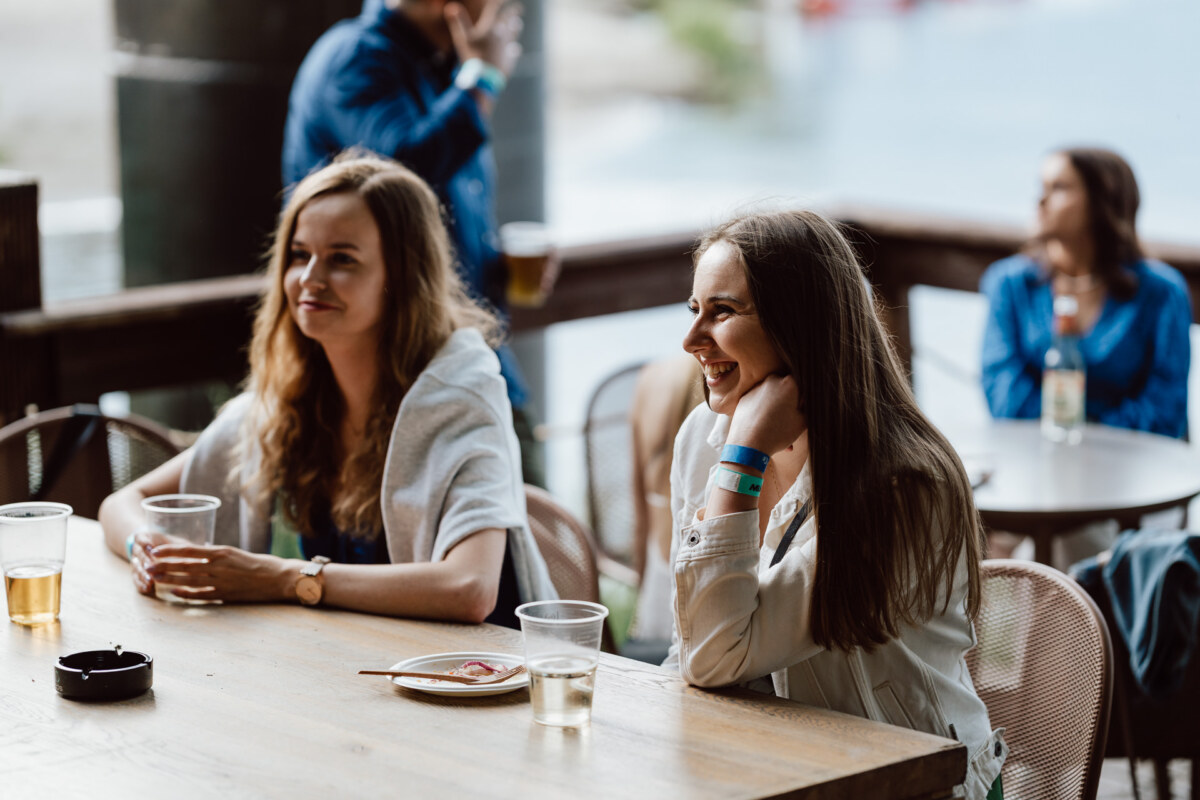 Two women sit at an outdoor wooden table, smiling and sipping drinks. Other people are visible in the background. The scene suggests a relaxed social gathering near the waterfront.  