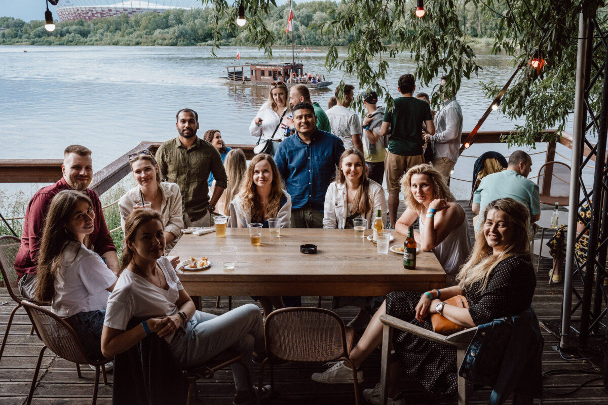 A group of people sit at a wooden table with drinks in the open air by the river. Trees provide shade, and a boat on the water can be seen in the background. The atmosphere is relaxed and sociable.  