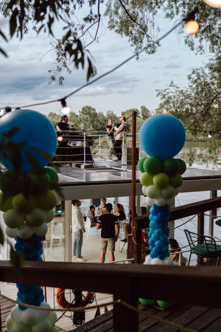 A group of people gather on a modern outdoor terrace near a lake, with blue and green balloon columns and string lights decorating the scene. Trees and water are visible in the background under a partly cloudy sky. 