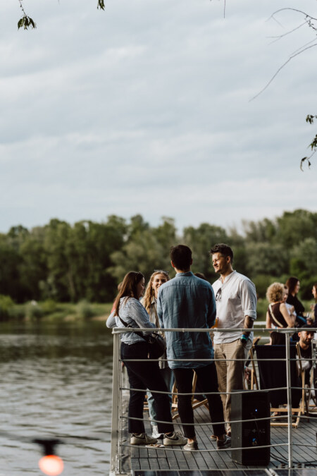 A group of people are standing and talking on a dock by the river, surrounded by trees and an overcast sky. Some are facing each other in conversation, while others are looking at the water. 