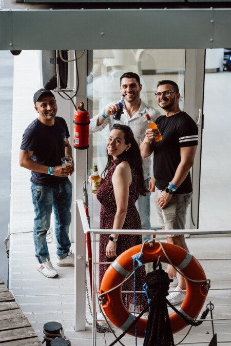 Four people are standing on the marina, smiling at the camera, each holding a drink. A lifebelt and fire extinguisher are visible nearby. The group seems to be enjoying a casual encounter near the water.  