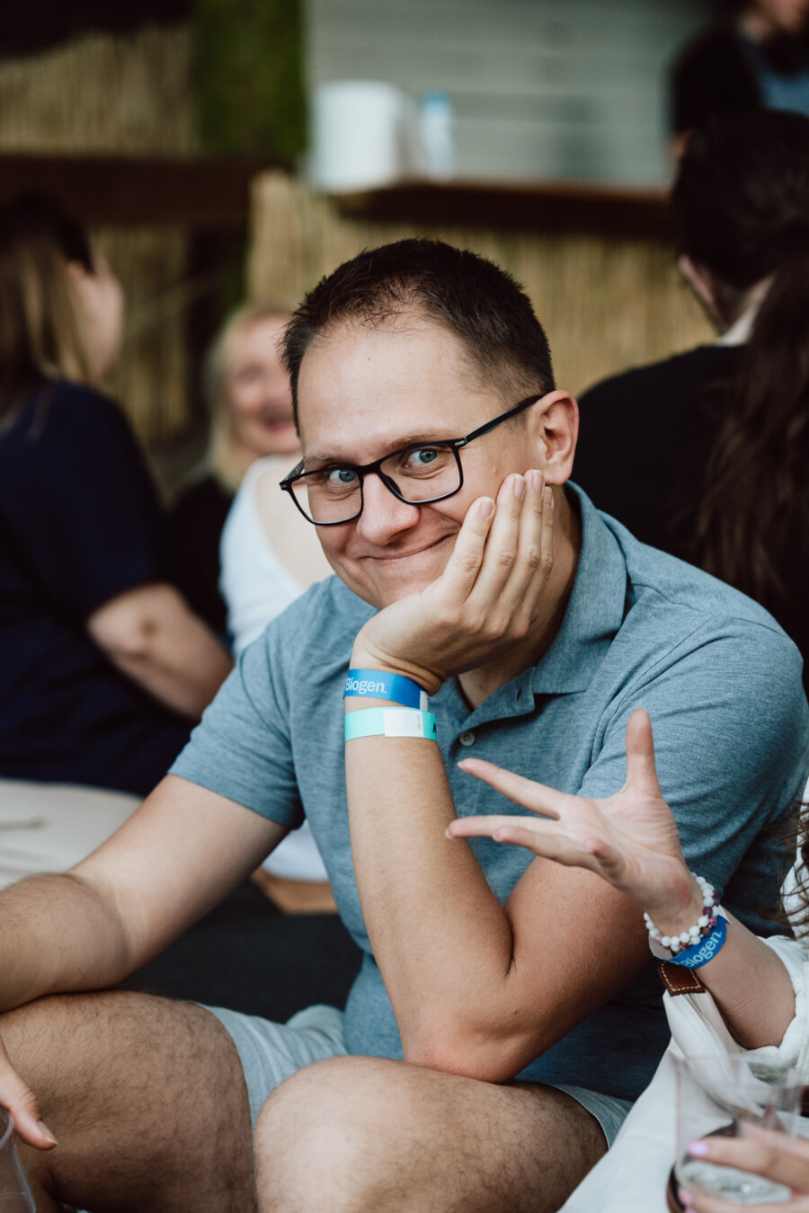 A man wearing glasses and a blue polo shirt sits with his chin resting on his hand, smiling at the camera. He has a blue event wristband on his wrist and is in a room, surrounded by other people. 
