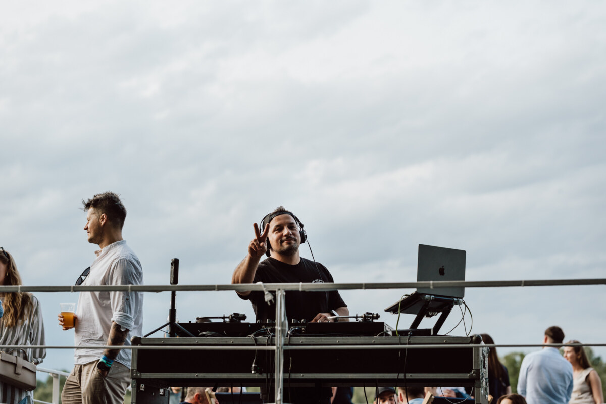 A DJ wearing headphones stands behind a booth with turntables and a laptop, smiling and making the peace sign. People gather outside under an overcast sky. 