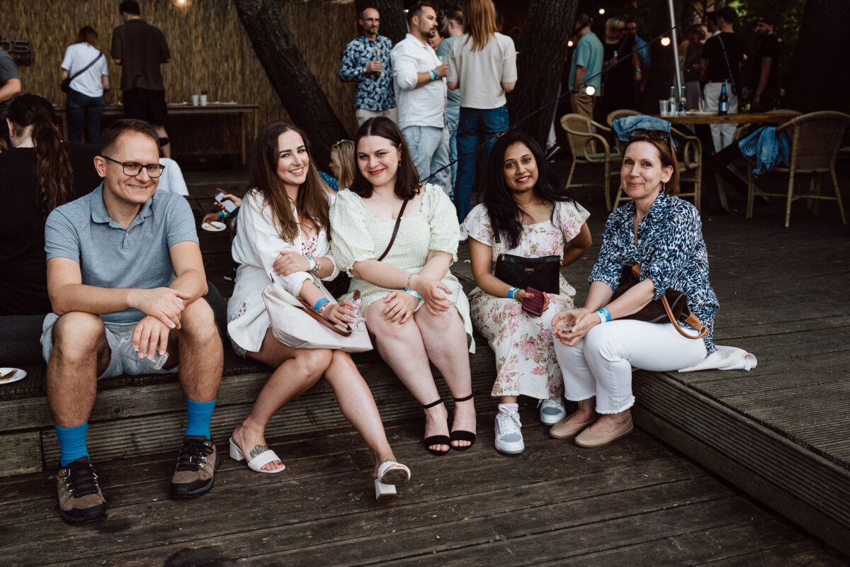 Five people sit and relax outdoors on a wooden terrace during a social gathering, smiling at the camera. Other people stand and mingle in the background near tables and trees decorated with string lights. 