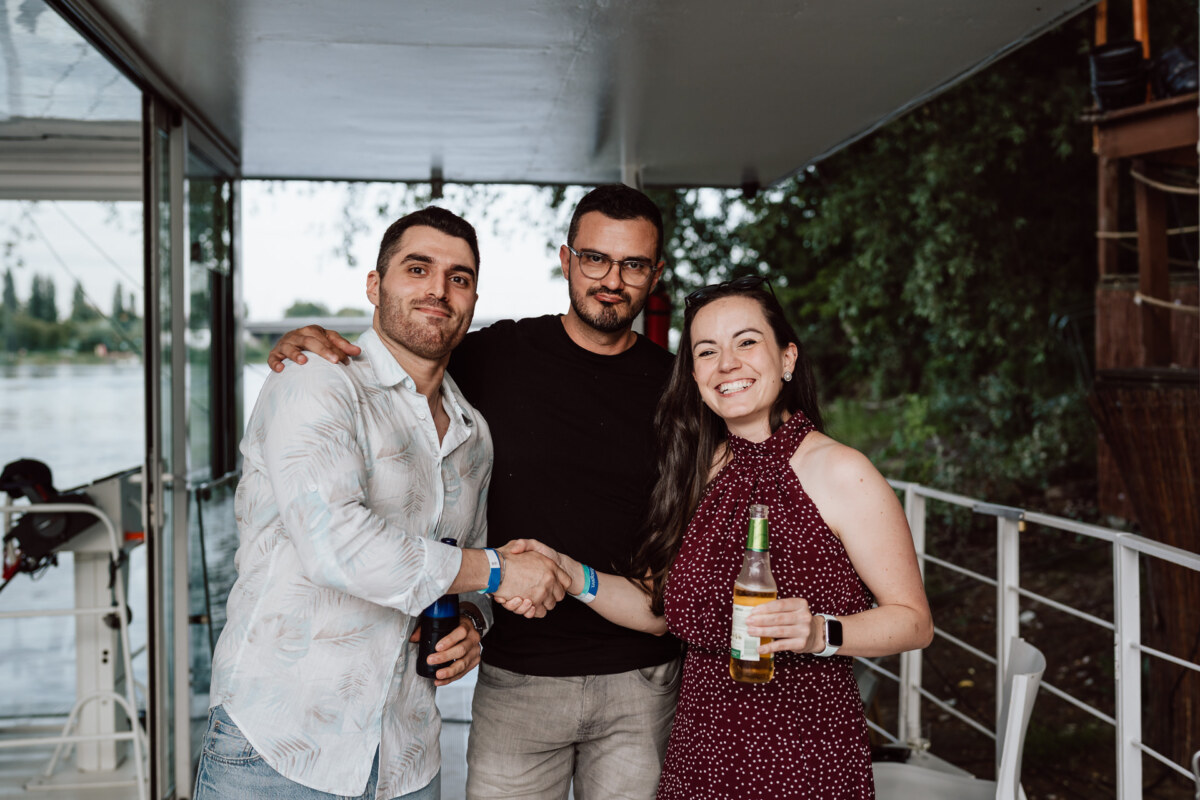 Three people are standing on a boat, two men are shaking hands and a woman is smiling while holding a drink. They look relaxed and are dressed casually, with trees and water visible in the background. 