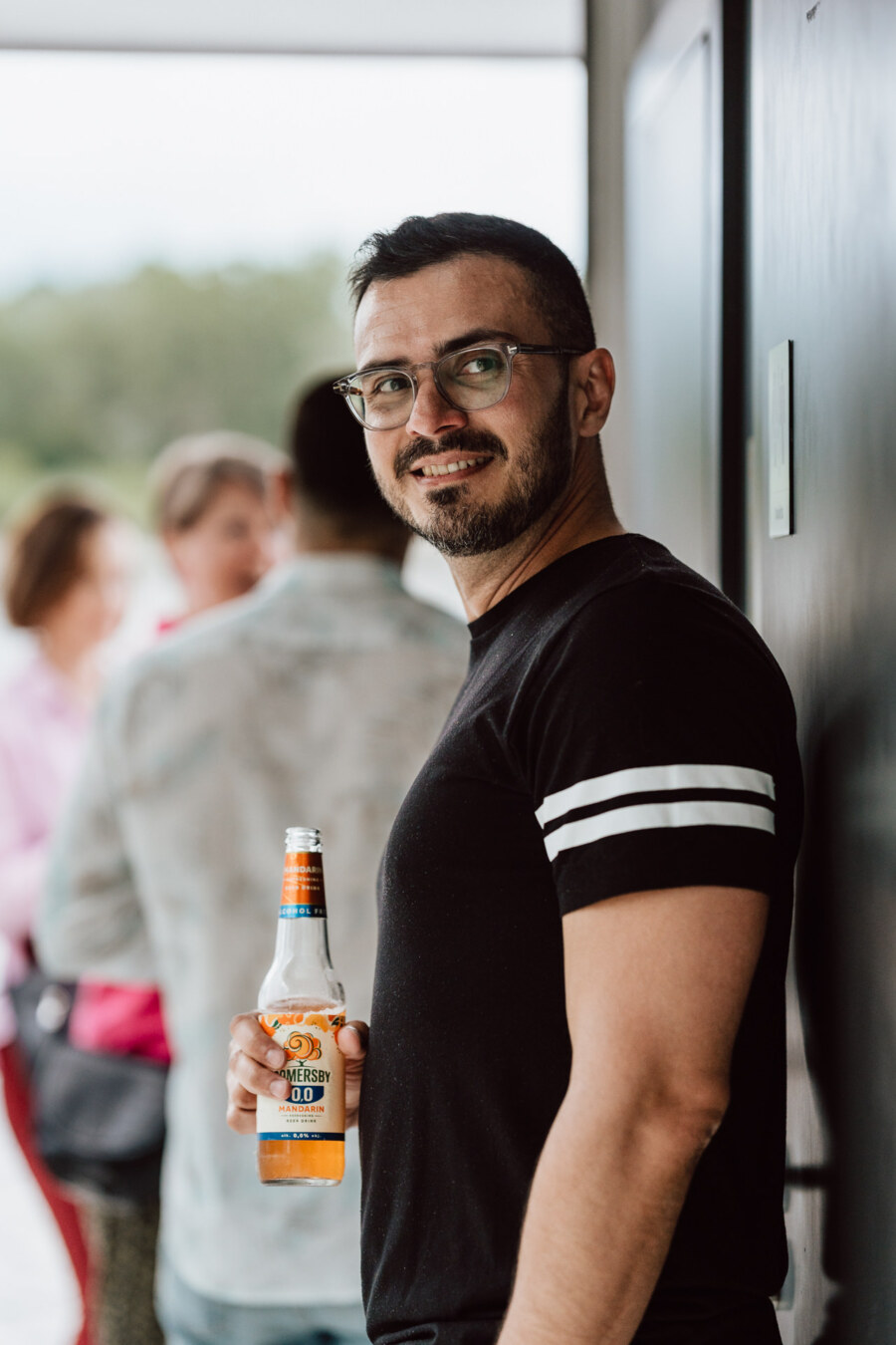 A man with glasses and a beard, wearing a black T-shirt with white stripes on the sleeves, stands in a room holding a bottle of non-alcoholic beer and smiling at the camera; there are indistinct people in the background.