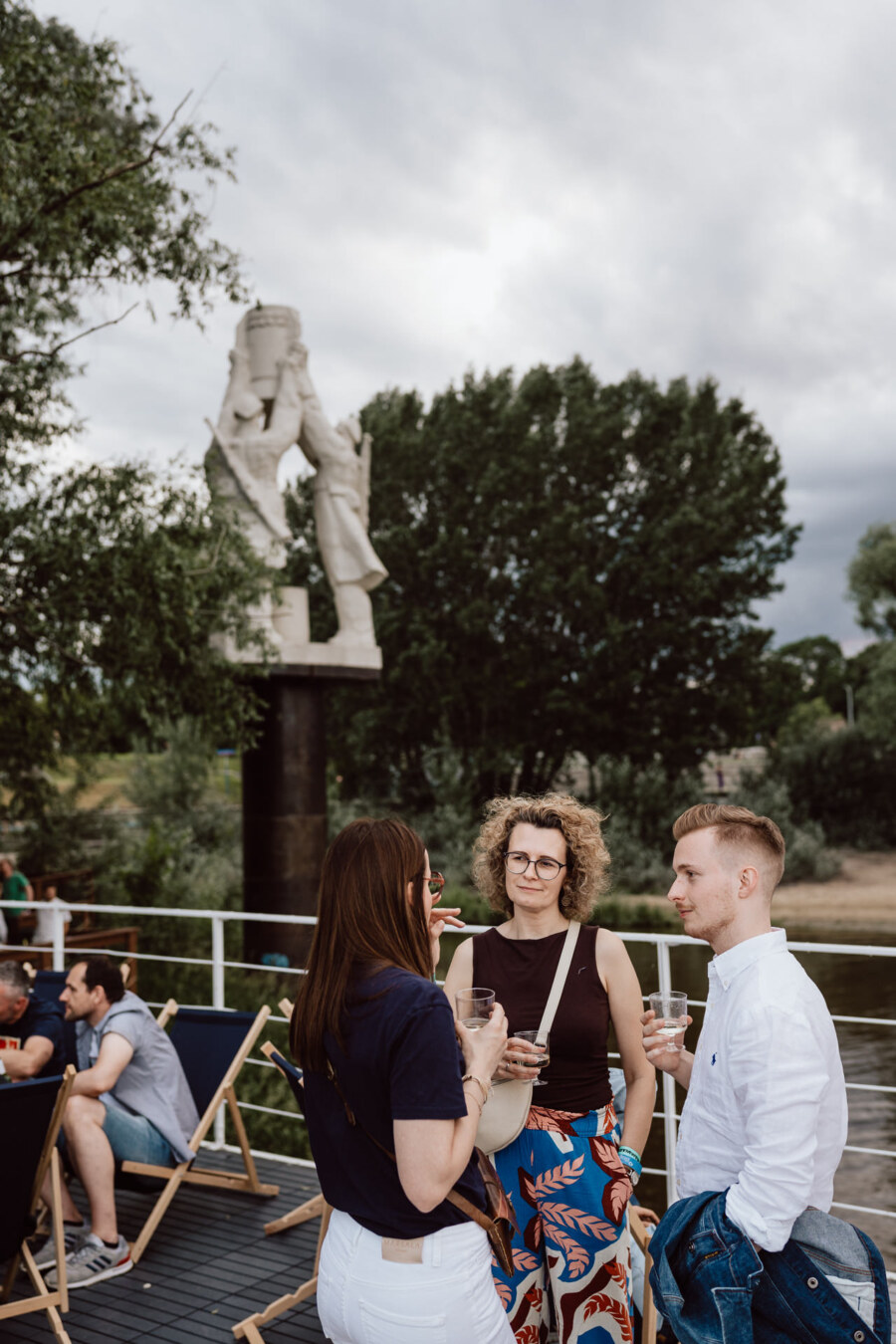 Three people stand and talk while holding drinks on an outdoor terrace with loungers. In the background is a large modern sculpture and lush green trees under an overcast sky. 