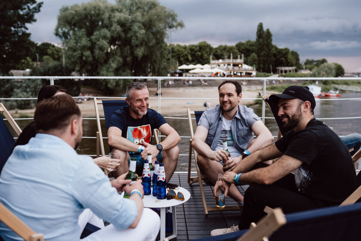 Five men sit on deck chairs near the river, smiling and chatting, holding drinks. Trees and a riverside cafe are visible in the background under an overcast sky. 