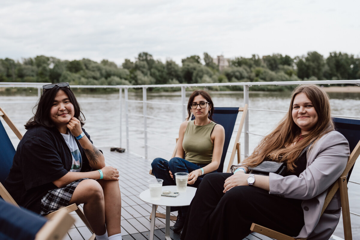Three people are seated in recliners around a small table with drinks, outdoors by the river. Trees and an overcast sky are visible in the background, creating a casual and relaxed atmosphere. 