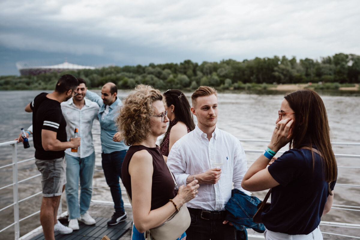 A group of people are standing and talking on the deck of a boat by the river, holding drinks and wearing casual summer clothes, with trees and an overcast sky in the background.