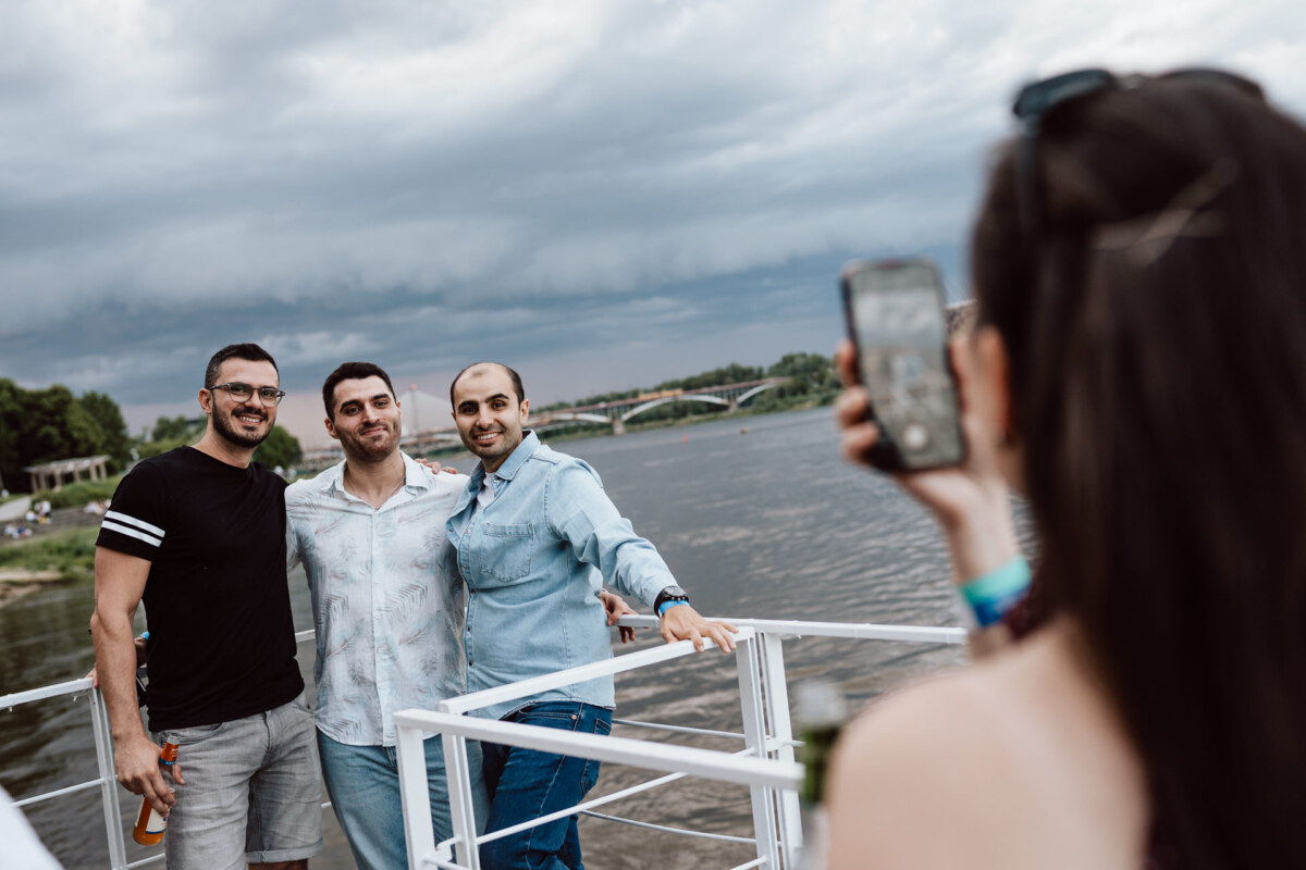 Three men stand smiling on a boat, posing for a photo. The woman in the foreground takes a picture of them with her phone. The river and bridge are visible in the background under an overcast sky.  