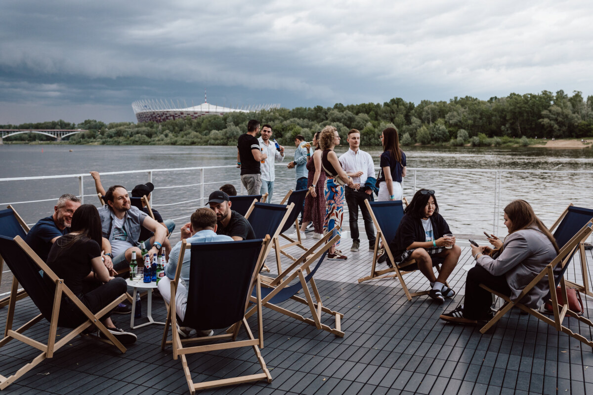 A group of people relax on deck chairs on a terrace by the river, with drinks on tables. The trees and stadium are visible across the river under an overcast sky. 