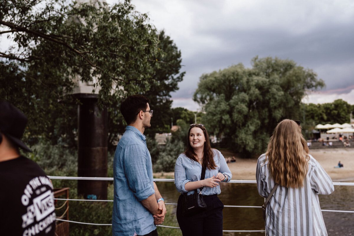 Three people are standing and talking by a river with trees and a sandy beach in the background. One person is turned away from the camera while the other two are smiling and talking. Overhead is an overcast sky.  