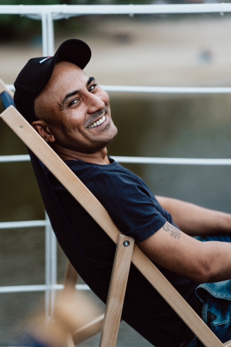 A man in a black cap and dark T-shirt sits relaxed on a wooden deck chair, smiling at the camera. He is outside near the water, with a blurry background of sand and greenery. 