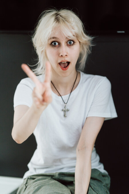 A young person with short, platinum blond hair and wide blue eyes, wearing a white T-shirt and a cross necklace, gestures a peace sign with her hand, looking surprised.