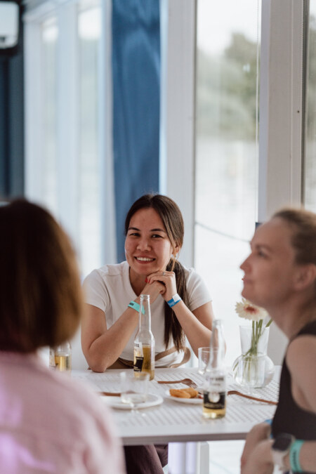 A woman smiles as she sits at a table with two other people, enjoying drinks and snacks in a bright, modern interior. Bottles and a vase of flowers are visible on the table. 
