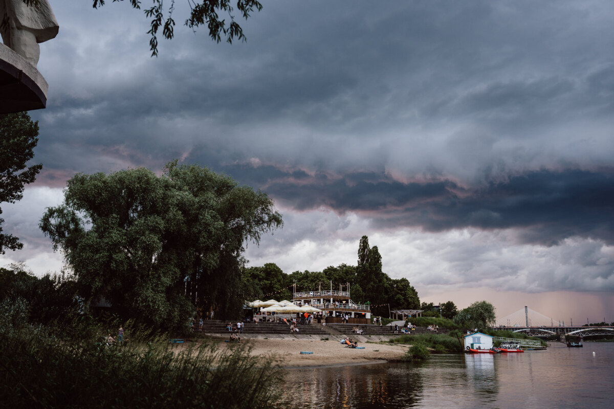 A sandy beach by the river with people gathered near the restaurant under large trees, with dark storm clouds overhead and a small boat docked by the water.