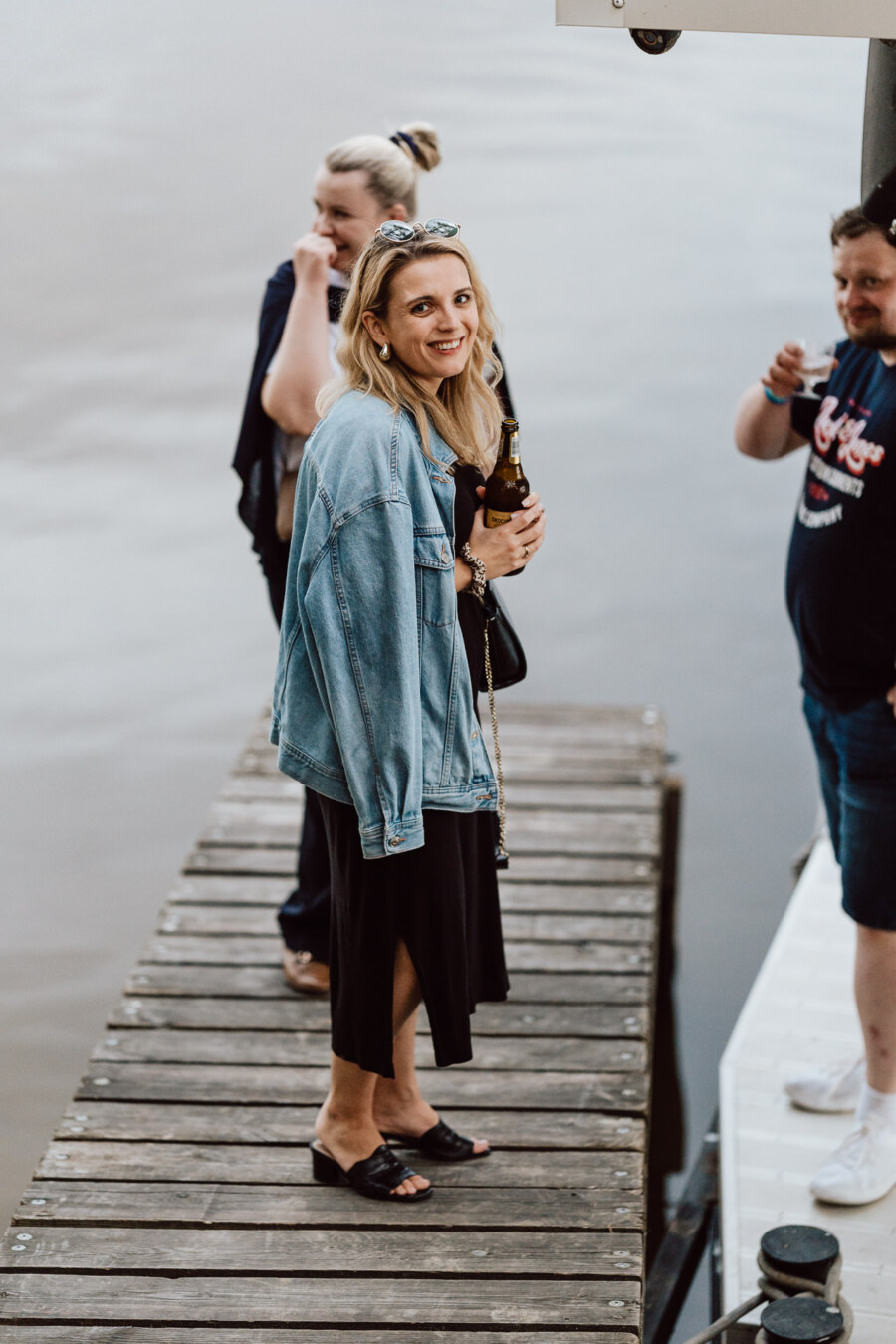 A woman in a denim jacket and black dress smiles as she stands on a wooden dock holding a drink. Nearby are two other people, one in the background and one on the right, both also holding drinks over the water. 