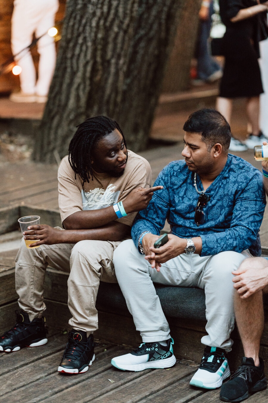 Two men sit on a bench outside and talk. One man gestures with his hand, pointing, holding a drink. Both are wearing casual clothes and sneakers, and trees and other people can be seen in the background.  