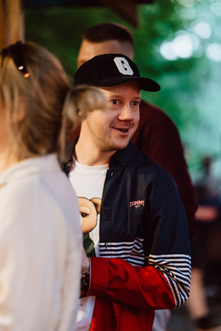 A man in a black cap and striped Tommy jacket smiles as he talks outside. A woman with sunglasses on her head is in the foreground, somewhat out of focus. The background is a fuzzy green.  