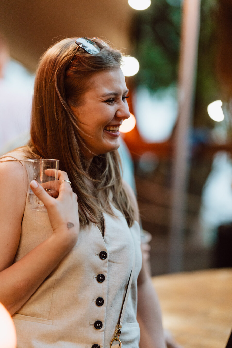A woman with long light brown hair, wearing a sleeveless beige top and sunglasses on her head, smiles as she holds a clear glass with a drink during a social gathering in the room.