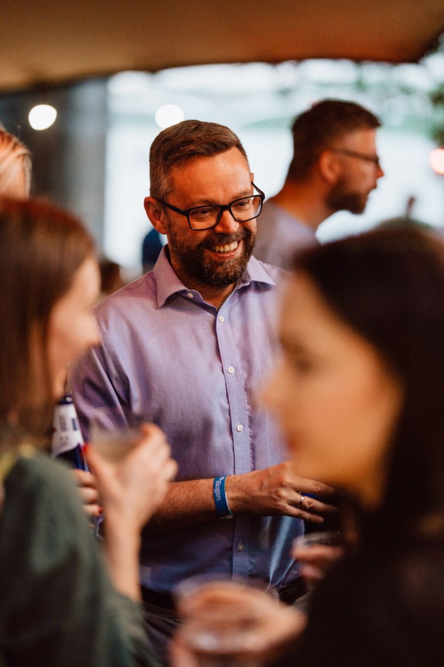 A man with glasses and a beard, wearing a light purple shirt, smiles while talking to others at a social gathering. In the foreground, blurry people holding drinks can be seen. 