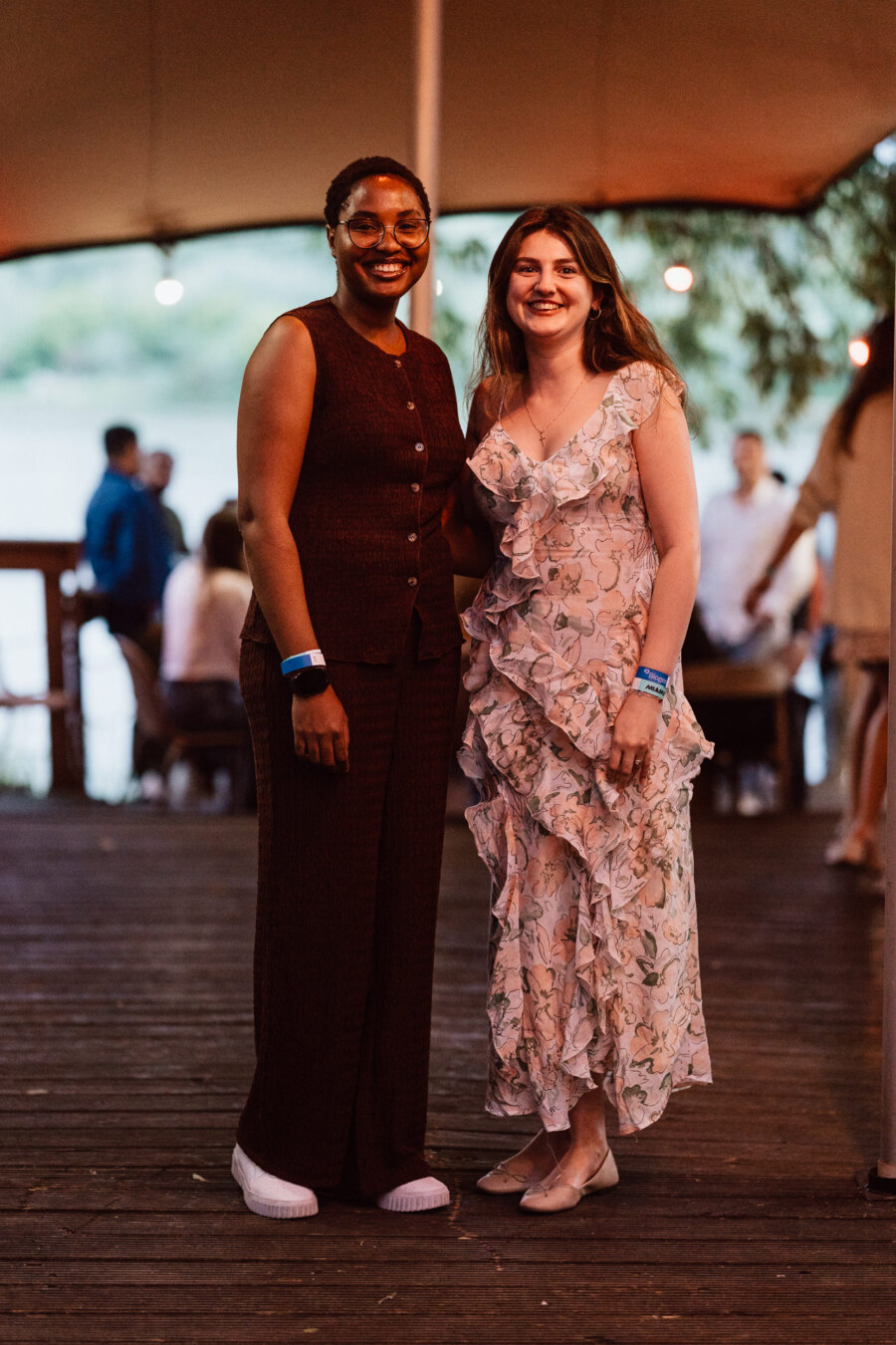 Two women are standing and smiling under a canopy. One is wearing a sleeveless brown outfit and glasses, while the other is wearing a light dress with floral ruffles. They are on a wooden terrace, with people and trees blurring in the background.  