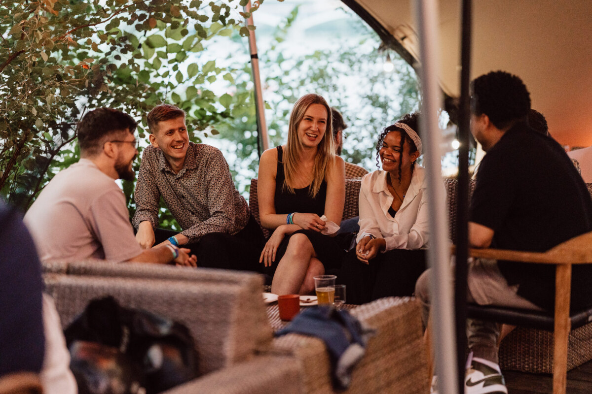 Five people sit together on outdoor wicker sofas, smiling and chatting. Drinks and cups stand on a table in front of them, while greenery and soft lighting create a relaxed, casual atmosphere. 