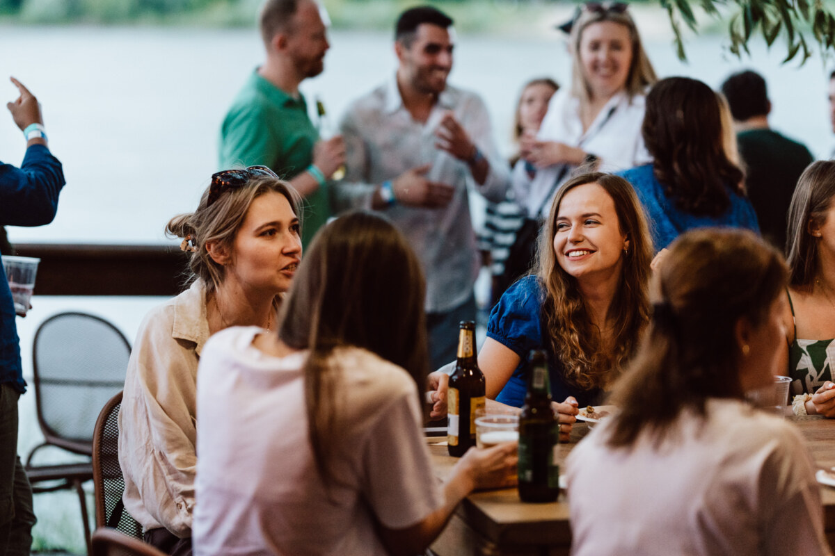 A group of people meet outdoors at a wooden table with drinks and food, smiling and chatting on a sunny day near a body of water. Some people stand in the background, mingling and having a good time. 