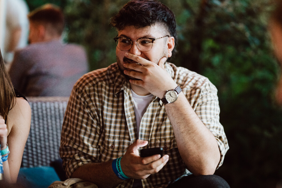 A man wearing glasses and a plaid shirt sits outside, holding a smartphone and covering his mouth with his hand, looking surprised or amused. The background is blurred with greenery and other people. 