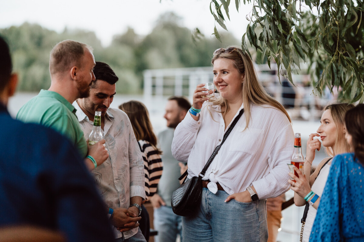 A group of people are standing together outside, socializing and smiling while holding drinks. Trees and fuzzy greenery are in the background, and everyone seems relaxed and engaged in conversation. 