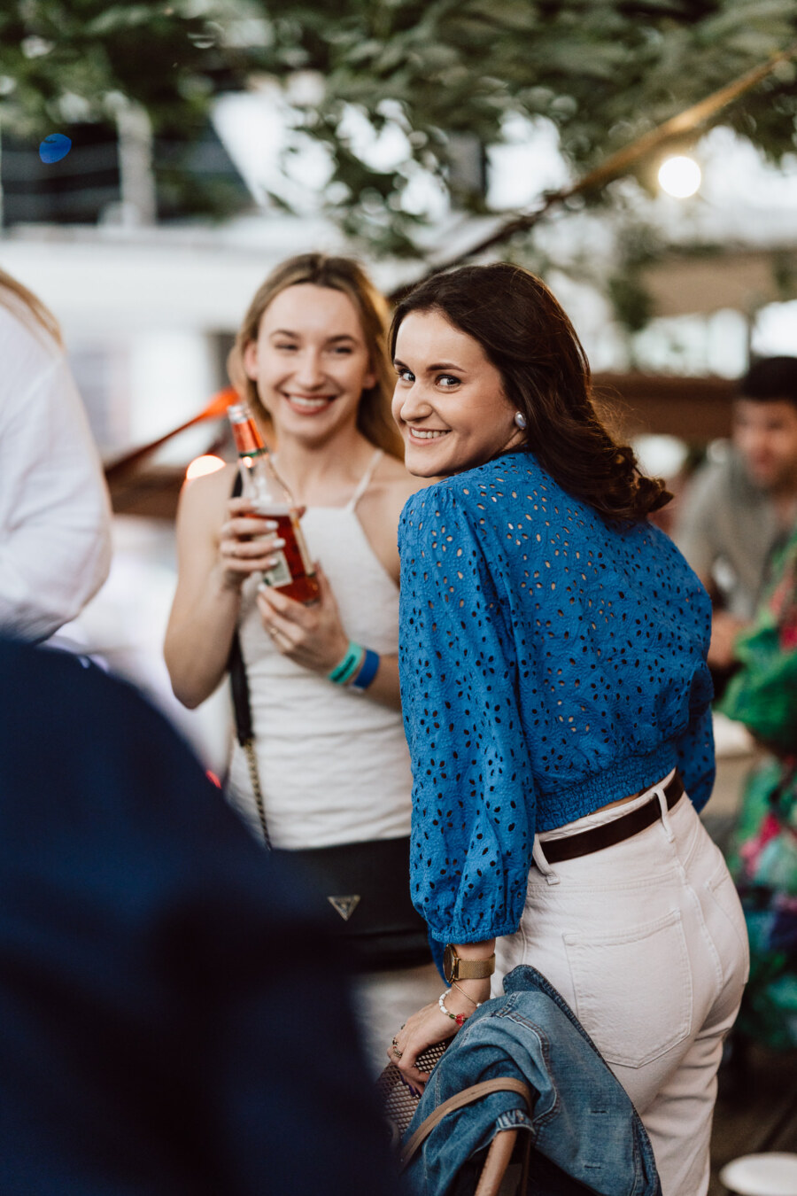 Two women are smiling at an outdoor party. One woman in a blue blouse and white pants looks over her shoulder while the other holds a drink and stands beside her, both looking happy and relaxed. 