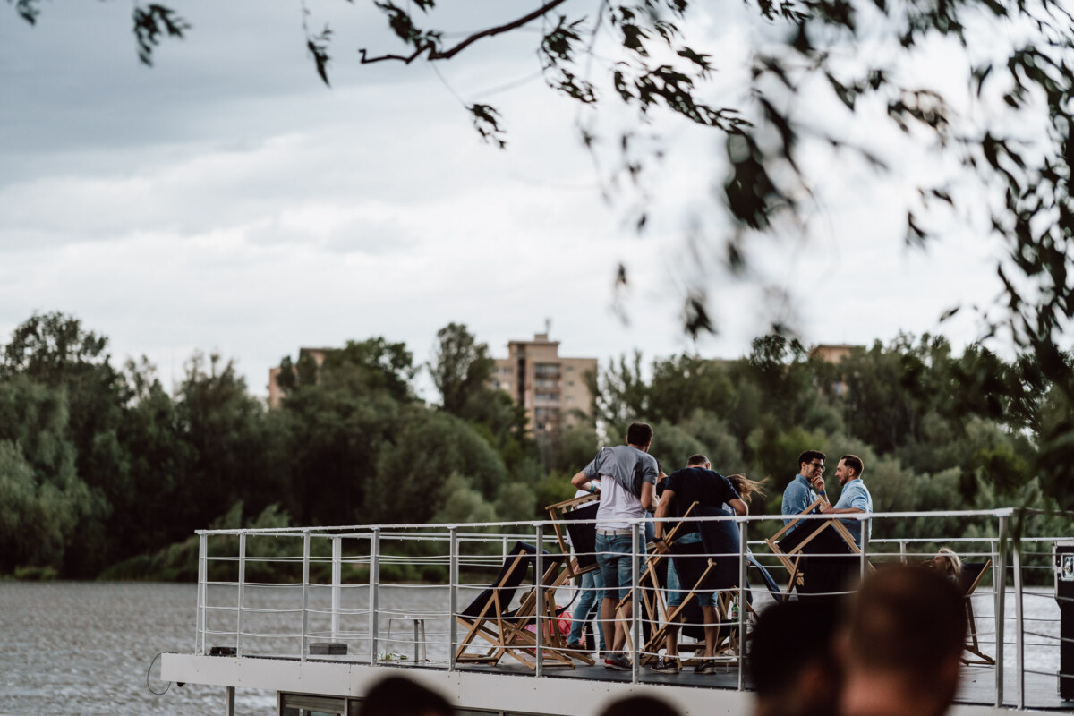 A group of people stand and sit on the deck of a boat on the river, surrounded by trees, with residential buildings visible in the background under an overcast sky.