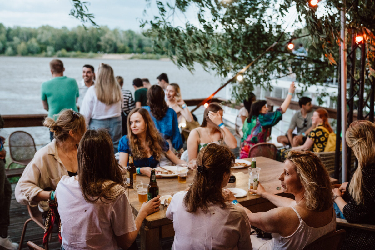A group of people gather outdoors at wooden tables near the river, enjoying drinks and conversation. Some sit, others stand, while trees and strings of lights create a festive, relaxing atmosphere. 