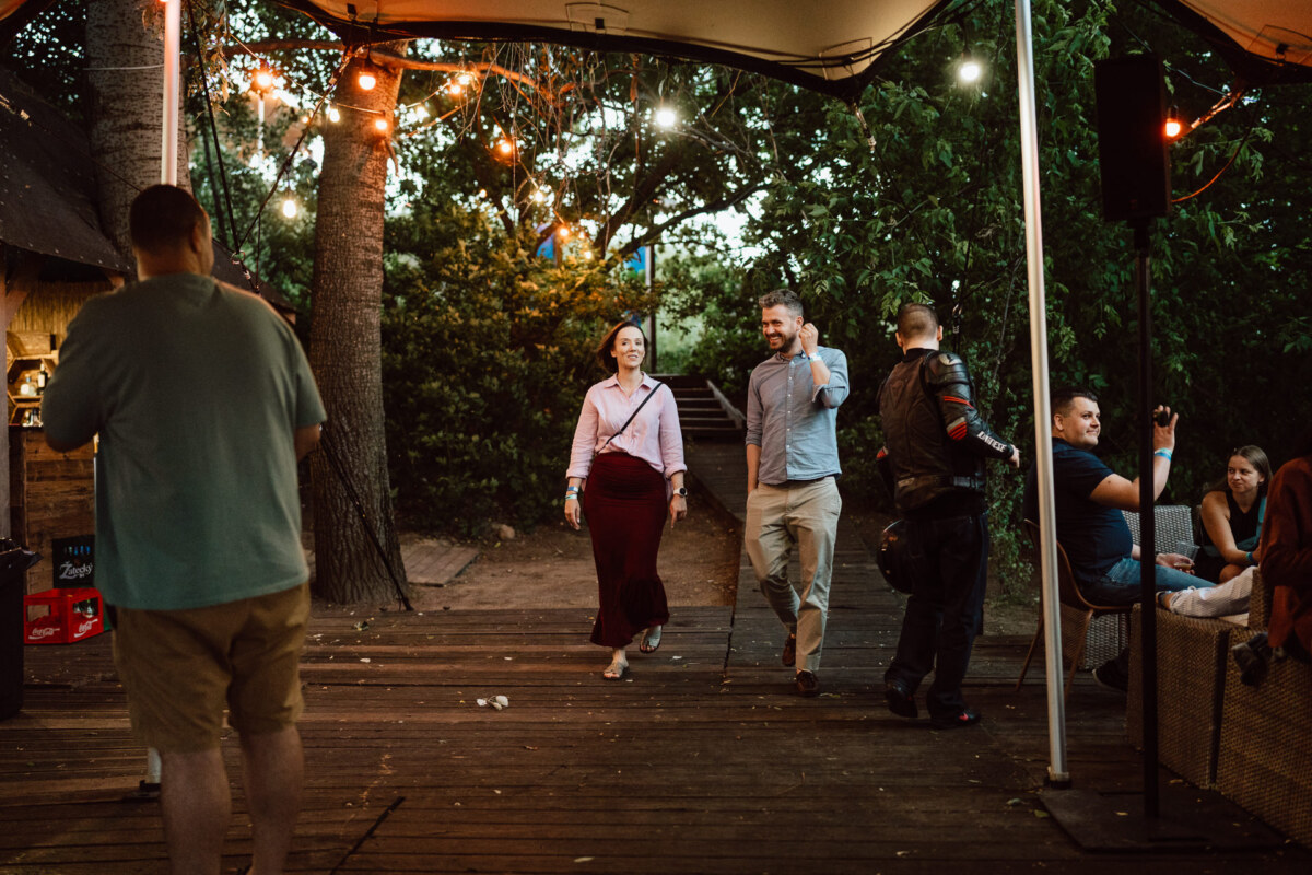 A man and woman walk together under string lights on a wooden terrace surrounded by trees, while people sit and stand nearby in a relaxing outdoor setting.