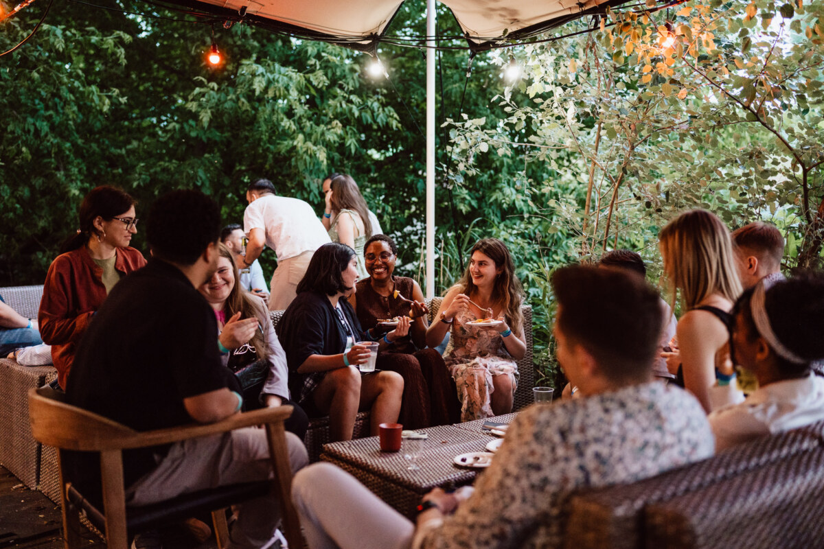 A group of people sit and talk in the open air under string lights, surrounded by greenery. Some are drinking and eating, creating a casual and joyful atmosphere. 