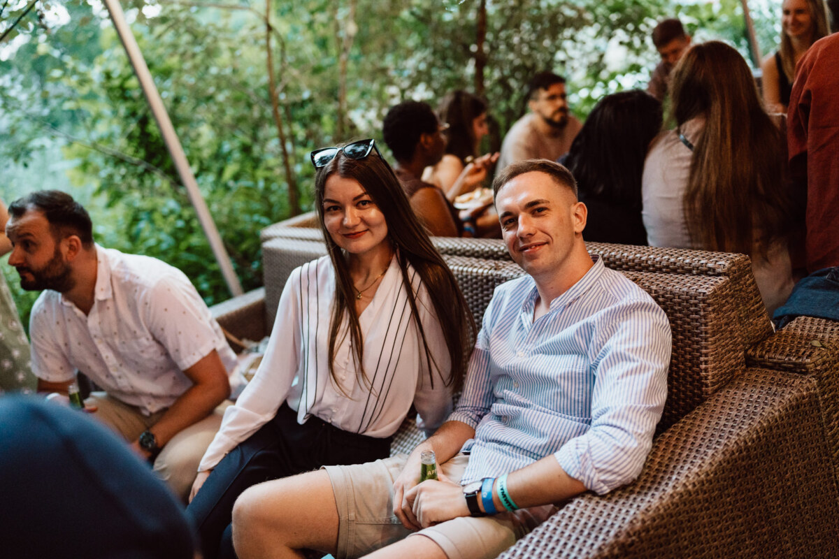 A group of people meet outside on plaid couches. A woman in a white blouse and a man in a striped shirt sit in the foreground, smiling at the camera. Others chat in the background among the greenery.  
