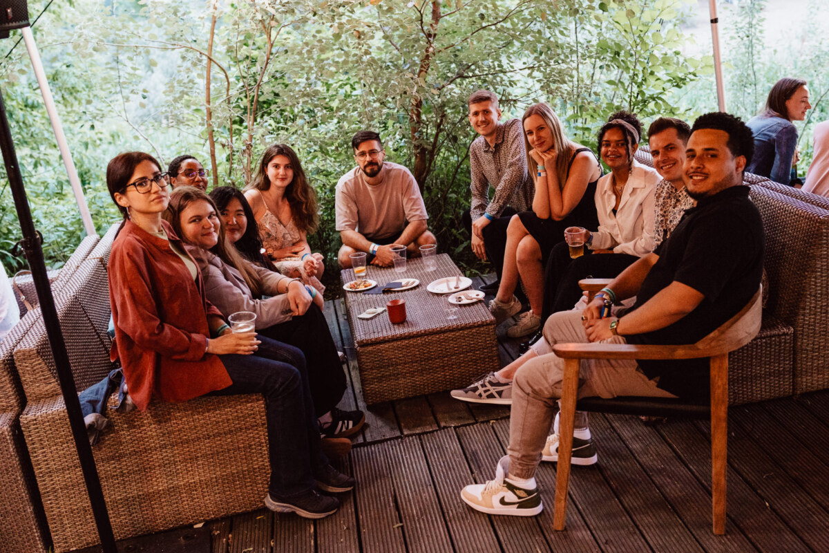 A group of people sit together on outdoor wicker furniture around a table with drinks and snacks, smiling and enjoying each other's company in the lush green garden.