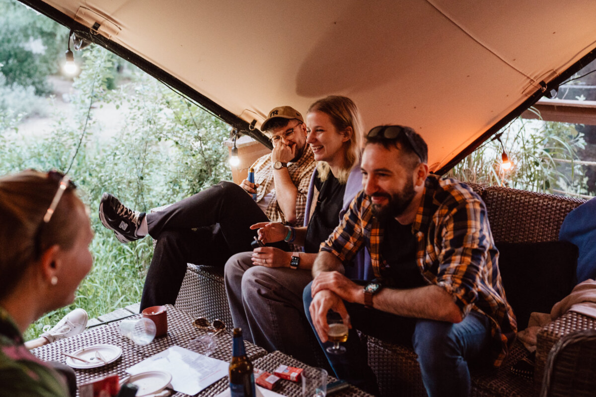 Four friends sit and laugh together in a cozy, covered outdoor space decorated with string lights. Drinks and snacks stand on a table in front of them, creating a relaxed, fun atmosphere. 