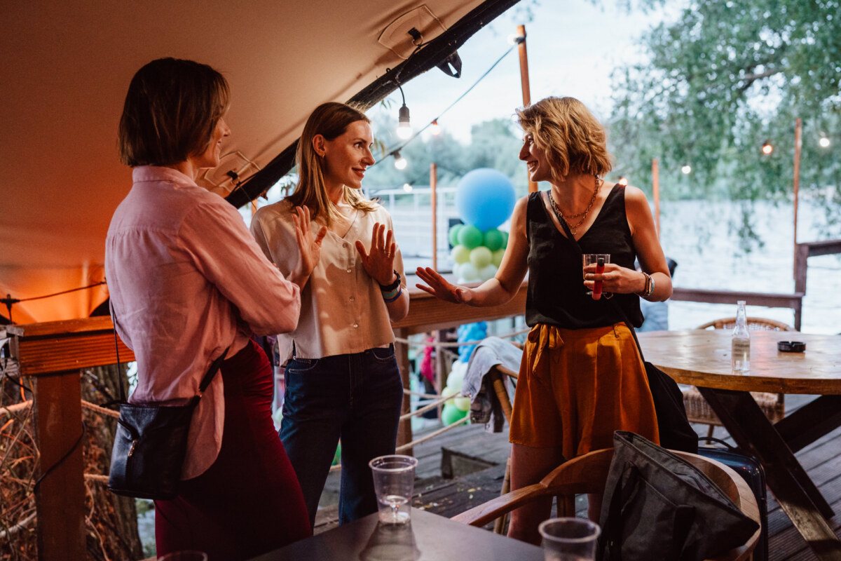 Three women stand and talk animatedly at an outdoor party near the water, with string lights, balloons and empty plastic cups on a wooden table nearby. One of the women holds a drink and gestures, smiling. 