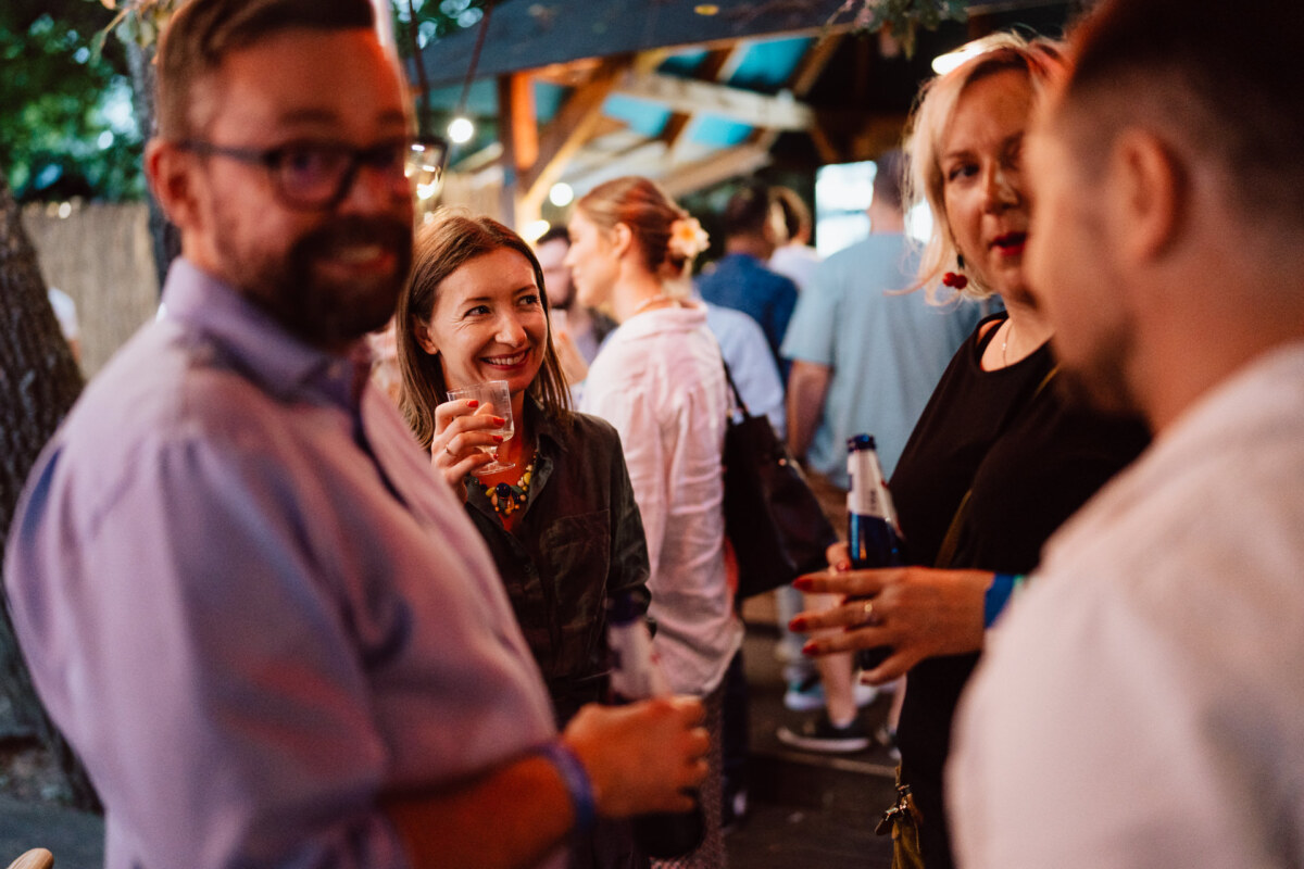 A group of people meet outdoors during a gathering, smiling and chatting while holding drinks. The atmosphere is warm and casual, with fuzzy lights in the background and guests mingling. 