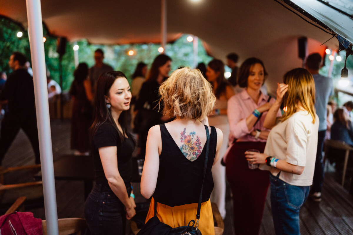 A group of people meet at an outdoor party under a canopy. In the foreground, a woman with a floral tattoo on her back is chatting with others. String lights and greenery create a festive, relaxing atmosphere.  