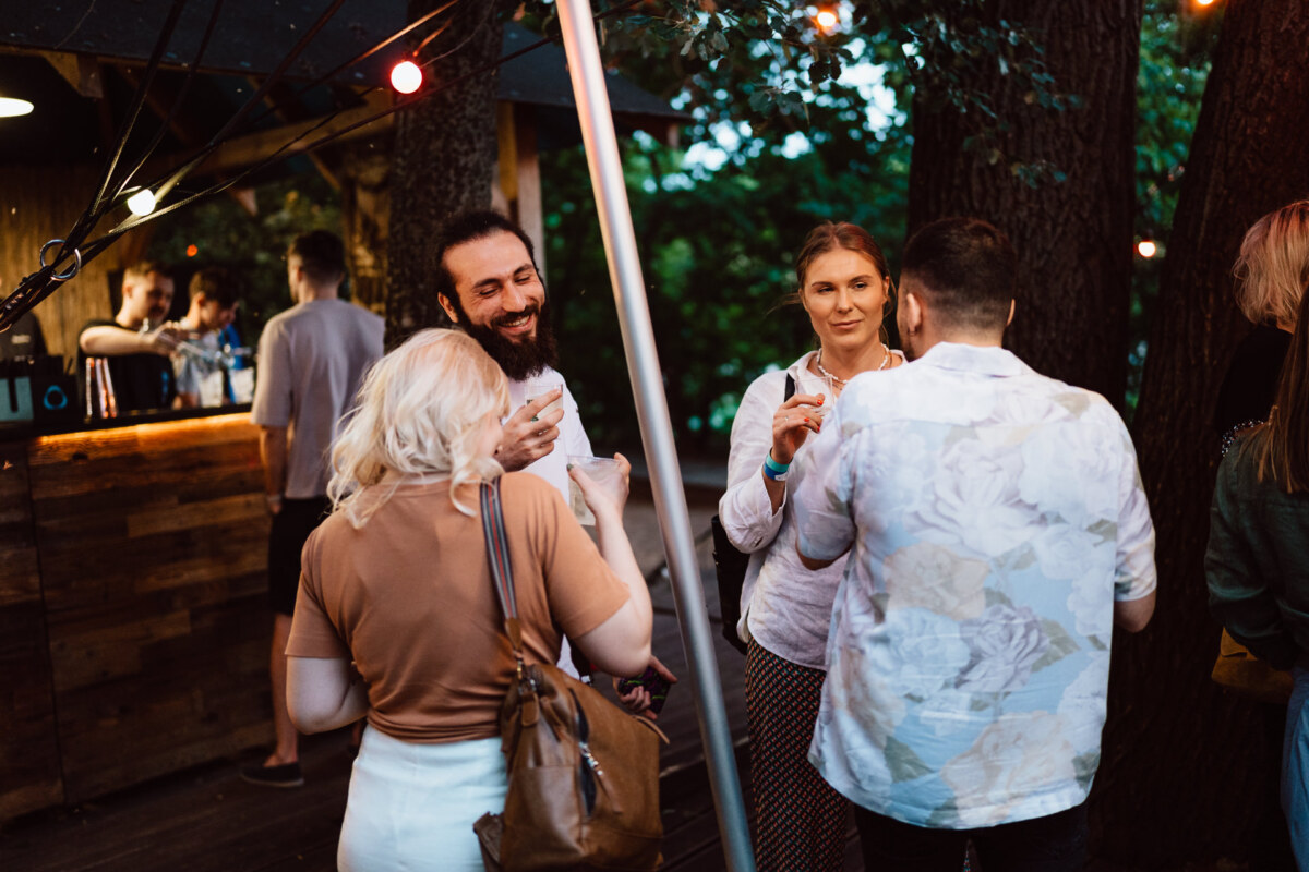 A small group of people meet outdoors in the evening near a wooden bar, surrounded by trees and string lights. Some hold drinks and talk, creating a casual and friendly atmosphere. 