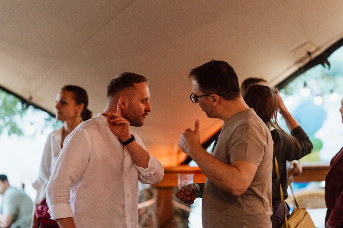 Two men talk at a social gathering, one gestures while holding a drink. Other people are talking in the background under a tent with warm lighting. 